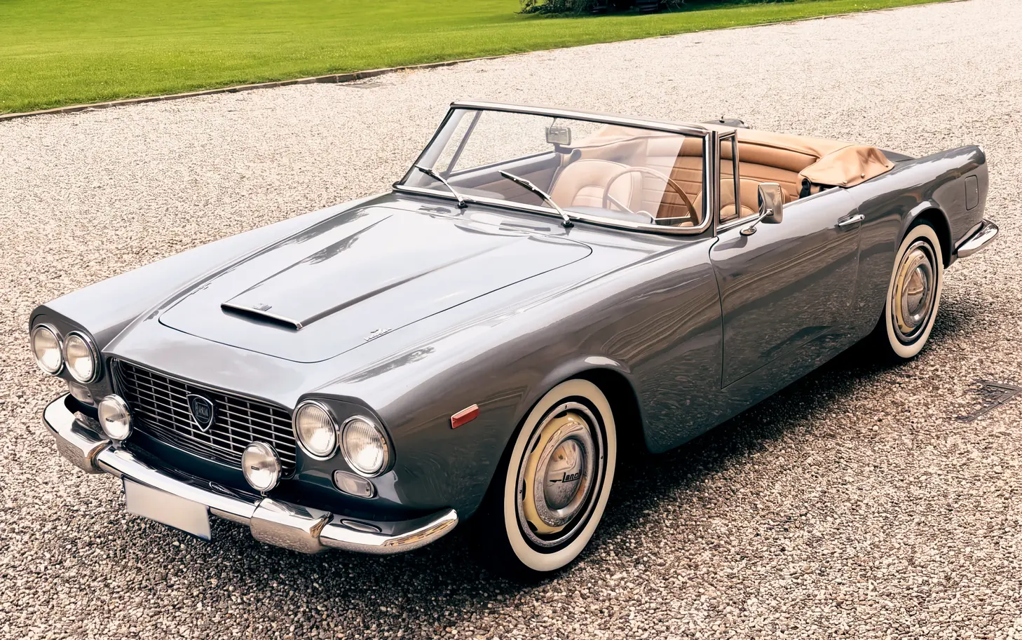 Una Lancia Flaminia cabriolet grigia con interni in pelle color cuoio, ferma su un viale di ghiaia accanto a un prato