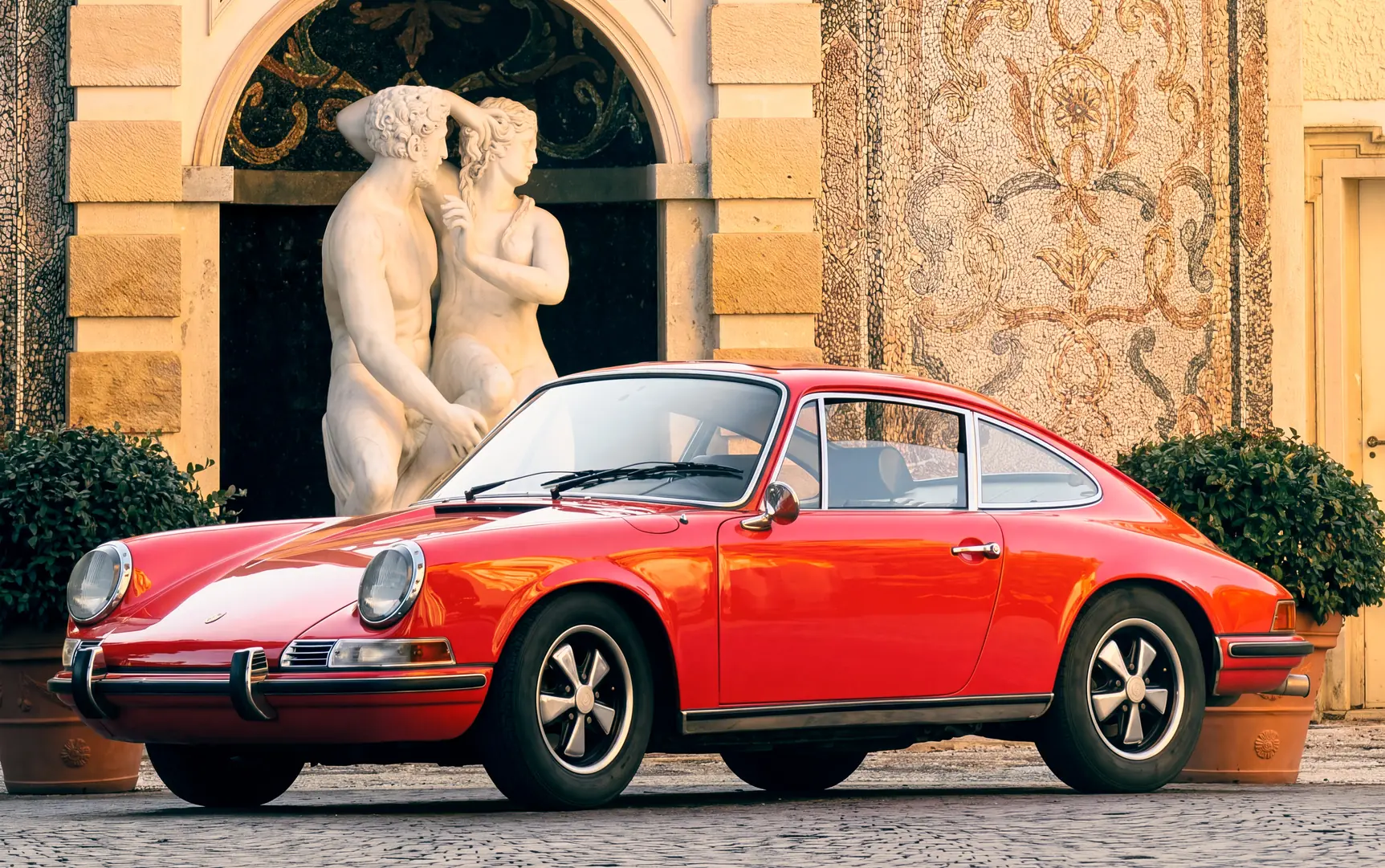 Una Porsche 911 T rossa del 1970 ferma in un cortile acciottolato, davanti a un gruppo scultoreo e a pareti a mosaico
