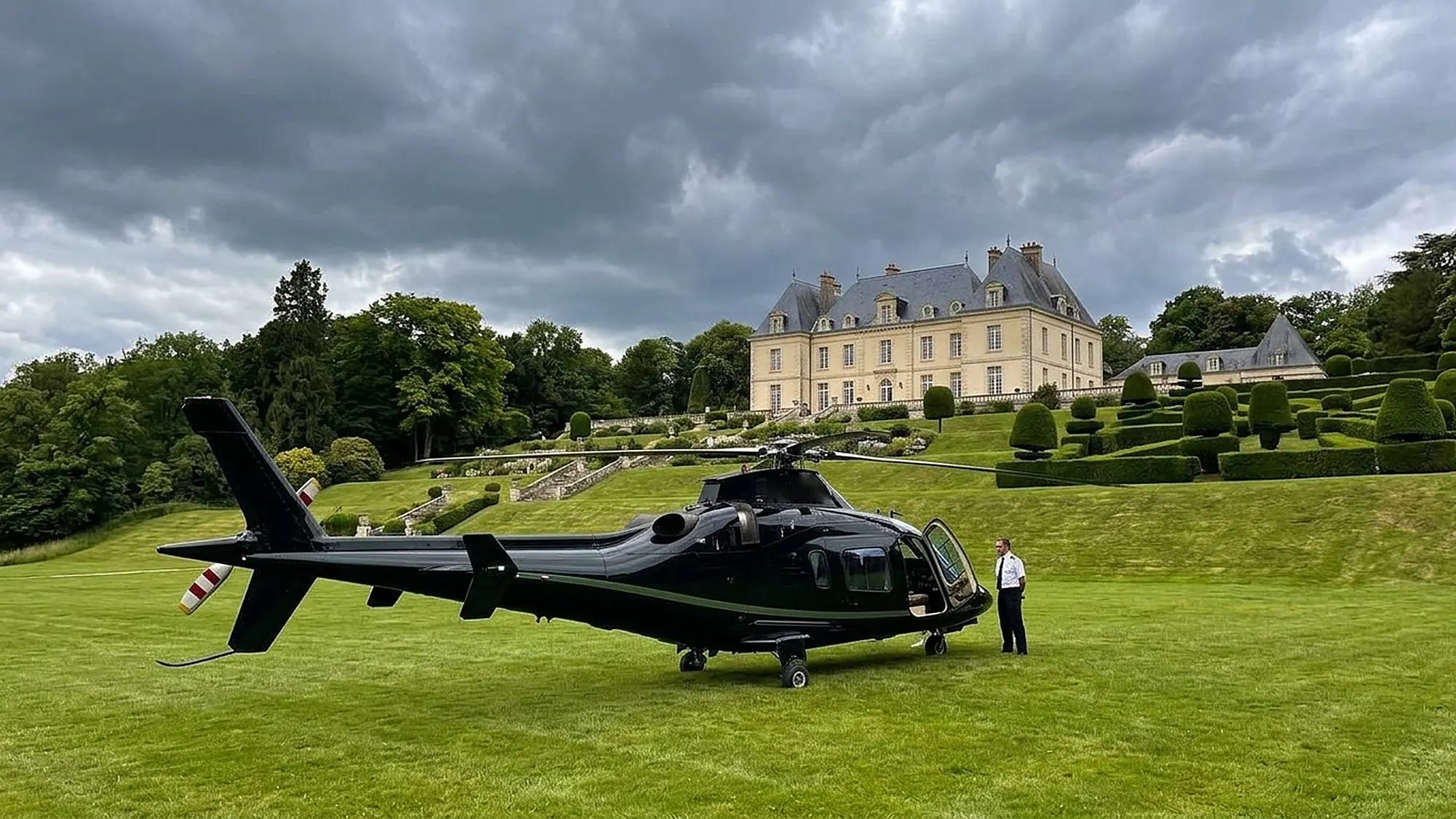 Elicottero nero posato sul prato di una villa storica con giardino di siepi scolpite, il pilota in attesa accanto alla porta aperta, cielo nuvoloso