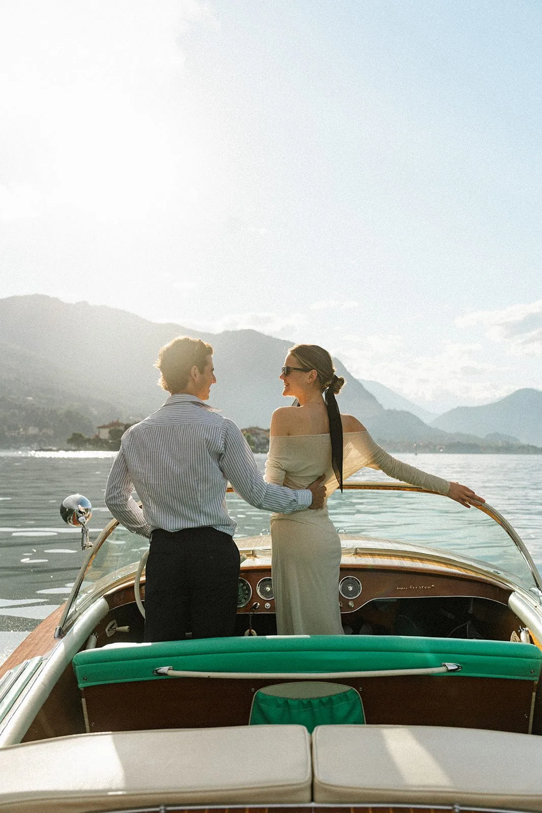 Una coppia in piedi a bordo di un Riva classico, di fronte al lago e alle montagne in controluce