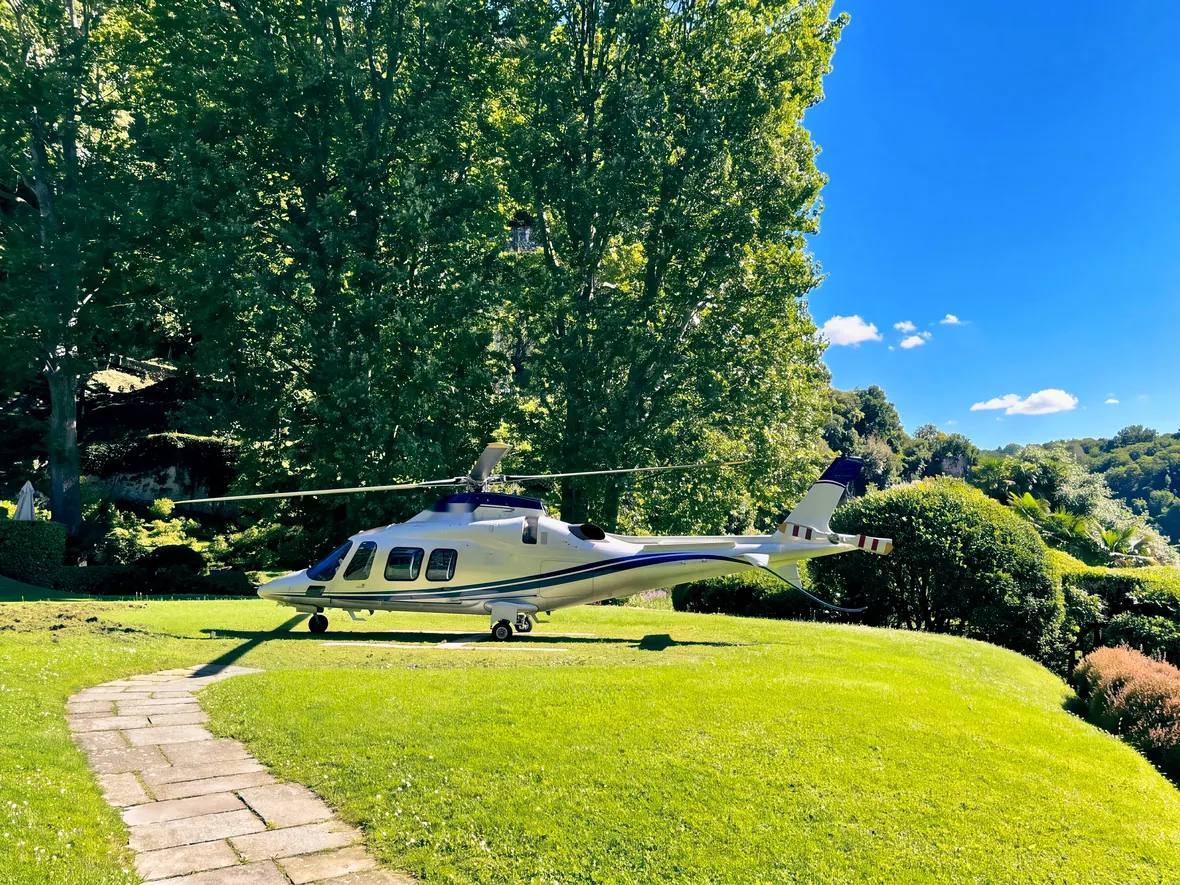 Un elicottero bianco posato sul prato di un giardino, fra gli alberi sopra il lago